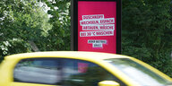 Ein gelbes Auto fährt vor einer rosa Werbetafel vorbei