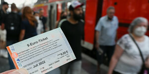 Eine Hand hält ein gedrucktes 9-Euro-Ticket ins Bild, dahinter sieht man unscharf einen belebten Bahnhof und einen Regionalzug der Deutschen Bahn.