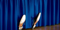 Aus einem nachtblauen Vorhang ragen zwei besohlte Schuhe heraus