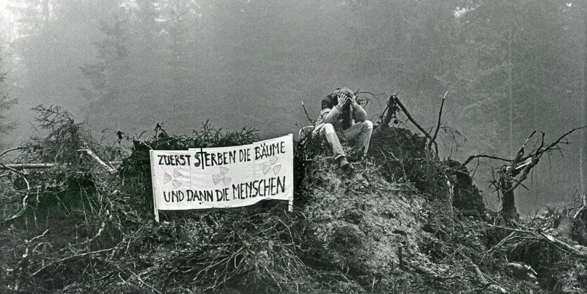 Ein junger Mann sitzt verzweifelt auf einem Berg abgestorbener Bäume. Vor ihm steht ein Transparent: Zuerst sterben die Bäume und dann die Menschen