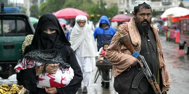 Ein Talib mit Gewehr steht auf einer Straße in Kabul, neben ihm geht eine verschleierte Frau