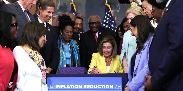 Nancy Pelosi unterzeichnet sitzend ein Dokument. Um sie herum stehen mehrere Abgeordnete der Demokraten. Sie freuen sich über die Verabschiedung des Gesetzes