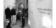 Schwarz-Weiß Bild. Drei Männer stehen vor einer Wand auf der steht: „Gerüchte verbreiten ist Landesverrat“