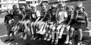 12 Kinder im Alter von ca. sechs Jahre sitzen auf einer Bank, im Hintergrund des Spielplatzes sind Mehrfamilienhäuser zu sehen