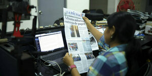 Eine Person sitzt vor einem Laptop und hält eine Zeitung in den Händen