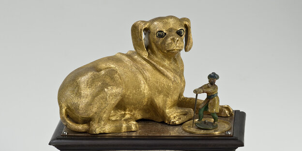 Auf einem Kasten sitzt ein Hund aus Metall, neben ihm kniet eine kleine Figur.