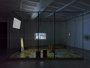 Ein Käfig, in dem sich vier Videomonitore befinden, sie zeigen unter anderem drei Vögel in einem Käfig auf einer Stange. Der Boden ist mit weißen und gelben Papierbögen ausgelegt, auf denen Schrift gedruckt ist. Mehrere Baumstümpfe stehen im Käfig