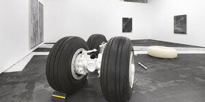 Blick in die Ausstellung von Daniel Hölzl. Im Vordergrund liegen Teile des Fahrwerks eines Flugzeugs auf dem Boden. Vor einem der Räder liegt ein Bremsblock. Dahinter ein aus Parafin nachgegossenes Rad. Weiße Bahnen überkreuzen den Boden.