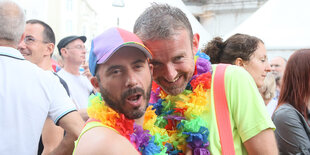 Zwei männlich gelesene Personen stehen in einer Menschenmasse und sind im Bildfokus. Zu sehen sind nur ihre Köpfe und Schulter. Sie lachen in die Kamera und haben beide Schmuck in Regenbogenfarben um dem Hals.