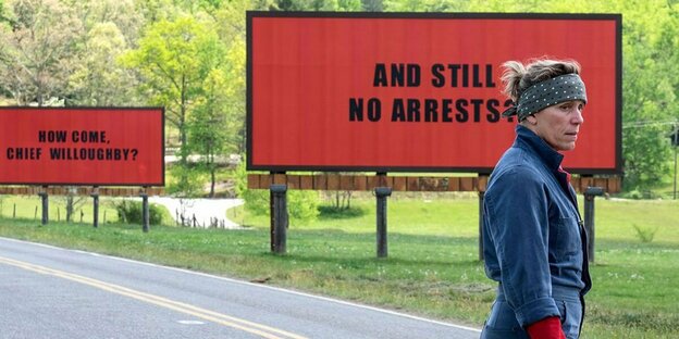 Szene aus „Three Billboards“: Mildred (gespielt von Francis McDormand) steht auf einer Landstraße, hinter ihr stehen zwei große rote Schilder mit schwarzer Schrift. Dort ist zu lesen „How Come, Chief Willoughby?“ und „And Still No Arrests?“
