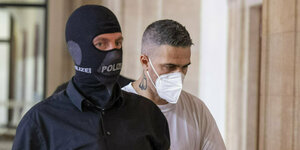 Ein maskierter Polizist gibt Bushido mit Maske und weißem Shirt Geleitschutz im Gericht