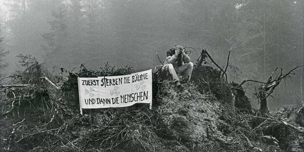Schwarzweißbild: Ein Demonstrant sitzt in geschädigtem Wald, neben ihm ein Transparent, Aufschrift: Zuerst sterben die Bäume und dann die Menschen