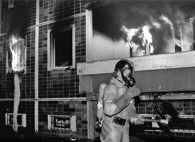 Eine Person in Gasmaske läuft an einem brennenden Gebäude entlang Eine Person in Gasmaske läuft an einem brennenden Gebäude entlang