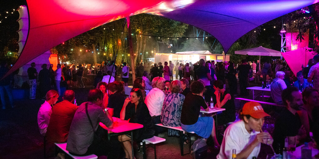 Viele Menschen sitzen nachts bei farbenfroher Beleuchtung auf Bierbänken und trinken.