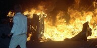 Ein Mann in einem weißen Jogginanzug steht nachts vor brennenden Autos