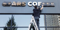 Ein Mann steht auf einer Leiter und entfernt die Schutzfolie von "Stars Coffee" an der Fassade