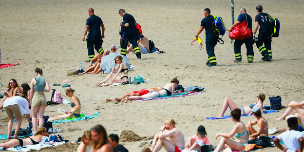 People lie on the beach in the heat, behind them a squad of firefighters with first-aid equipment