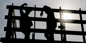 Zwei Arbeiter stehen auf einem Gerüst auf einer Baustelle und unterhalten sich. Im Hintergrund ist die Sonne zu sehen
