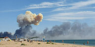 Bild eines Strandes mit Badegästen, im Hintergrund steigt eine riesige Rauchsäule in den Himmel