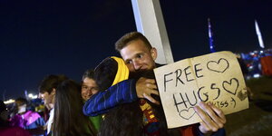 Ein junger Mann hält ein Schild mit der Aufschrift "Free Hugs" und umarmt eine junge Frau.