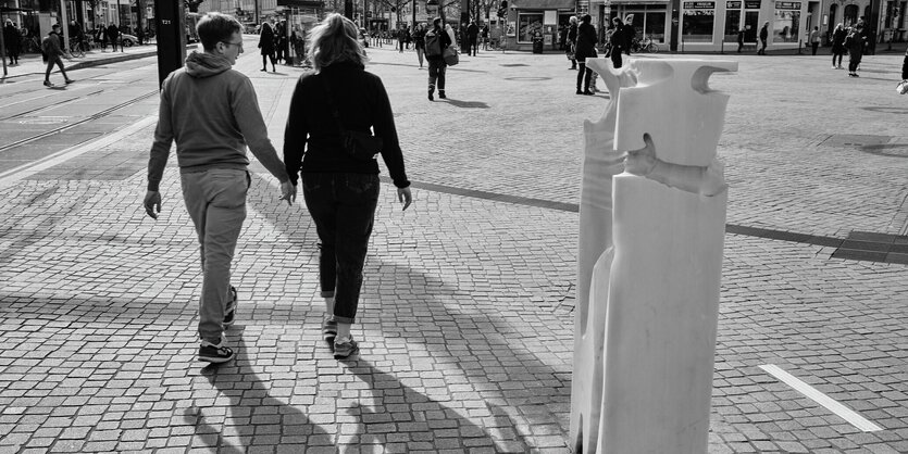 Zwei Personen laufen auf einem Platz. Sie halten Händchen. Rechts neben ihnen im Bildvordergrund eine viereckige Säule aus stein. In ihre sind Stellen freigelassen worden, die eine Umarmung zeigen.