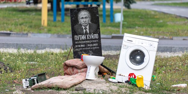Schwarzer Grabstein mit Putin-Gesicht, eine Closchüssel, eine Waschmaschine, Kinderspielsachen und Kissen auf einem Boden vertstreut