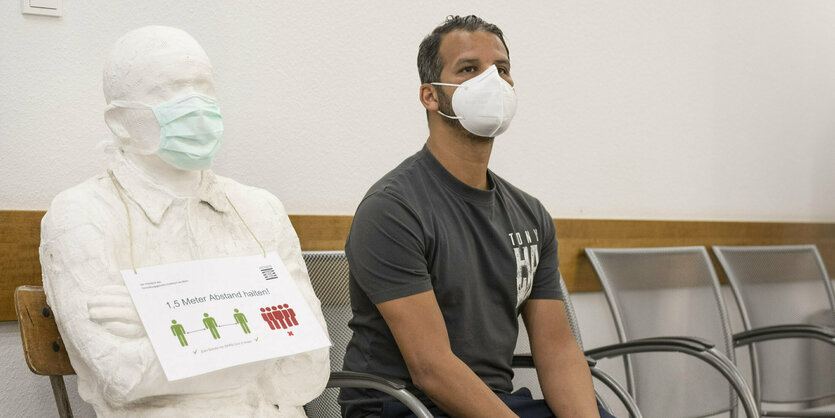 Abdelkarim Sedjerari sitzt mit Maske auf einer Bank, neben ihm eine Figur mit Schild, die darauf hinweis, dass 1,5 Meter Abstand gehalten werden soll