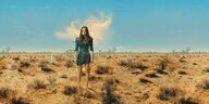 Serienbild. Eine Frau mit langen braunen Haaren steht im kurzen blauen Kleid in der Wüste.