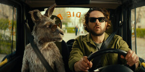 Ein Mann mit Bart und Sonnenbrille sitzt am Steuer eines Autos, neben ihm ein Känguru, ebenfalls mit Sonnenbrille