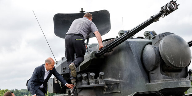 Kanzler Scholz und ein weiterer Mann klettern auf ein Panzerfahrzeug, auf dem Turm eine große Radarschüssel