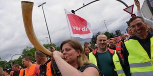 Beim Streik der Hafenarbeiter*innen bläst eine Frau im Blaumann in eine Art Horn. Hinter ihr stehen weitere Hafenarbeiter in Warnwesten, sie lachen. Verdi-Fahnen wehen