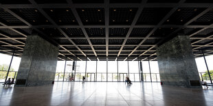 Eine Einganghsalle ist von innen im Weitwinkel fotografiert. Das Licht föllt von draußen herein. Die Halle ist fast komplett leer, nur zwei breite, graue Säulen stützen die Decke links und rechts. Die Decke hat horizontale und vertikale Querverstrebungen. Zwischen den Verstrebungen sind immer je vier kleine Lichter angebracht.