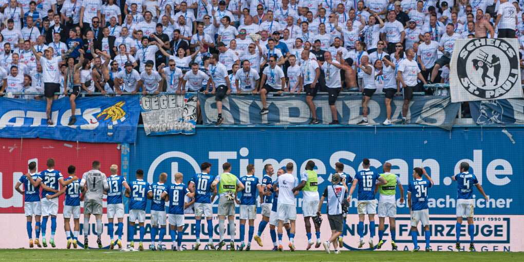 Die Mannschaft des FC Hansa Rostock steht vor der Tribüne mit jubelndem Fans, am Rand ist ein Transparent mit der Aufschrift "Scheiß St. Paul" zu sehen