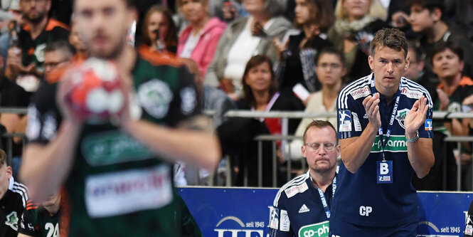 Im Vordergrund fängt ein Spieler den Ball, Christian Prokop steht im Hintergrund und feuert das Team an