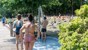 Eine Person mit Badehose steht unter einer Dusche im Freibad. Eine Person im Badeanzug und eine im Bikini warten davor. Im Hintergrund liegen Menschen auf einer Wiese und sonnen sich.