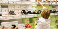 Eine Frau steht vor einem Regal mit Schuhkartons und Schuhen in einem Geschäft. Sie greift mit dem Arm nach einem Karton etwas über Kopfhöhe.