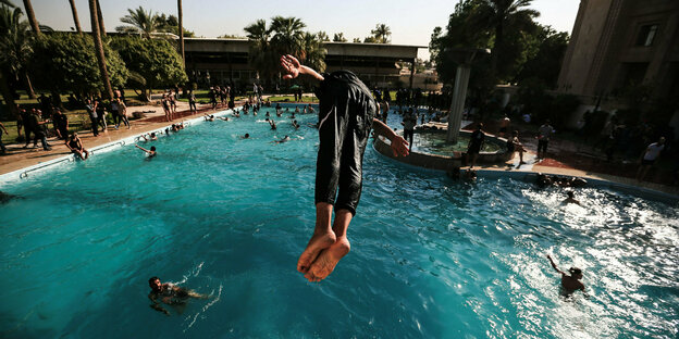 Ein großes Schwimmbecken mit vielen Menschen, im Hintergrund ein Garten und ein großes Gebäude. Vordergrund: Ein Mann macht einen Kopfsprung ins Wasser