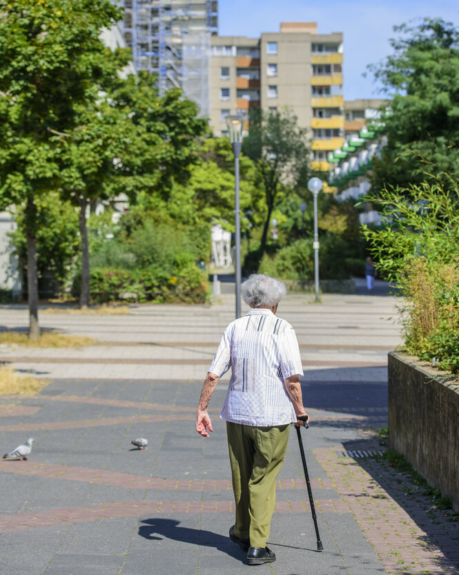 Eine alte Frau mit Stock entfernt sich, man sieht sie von hinten. Im HIntergrund Bäume und Plattenbauten