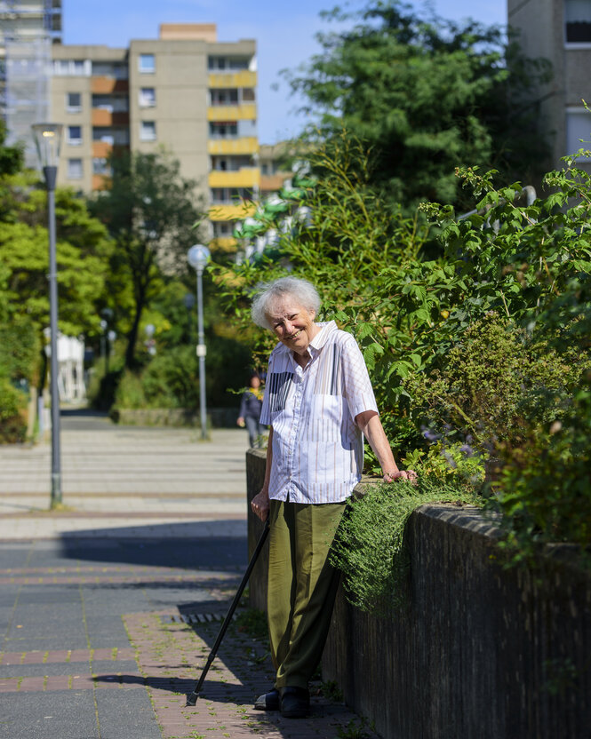 Eine alte Frau mit Stock steht an eine niedrige Mauer gelehnt und lacht. Hinter ihr Bäume und Plattenbauten.