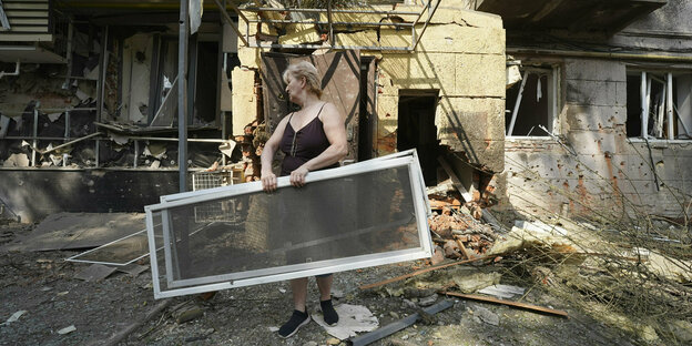 Eine Person mit zwei Fensterscheiben vor einem zerbombten Haus.