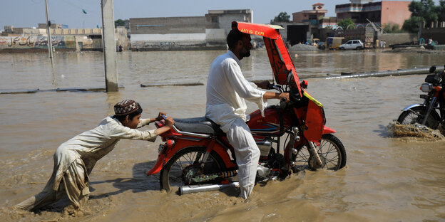 Eine Junge schiebt einen Mopedfahrer über einen überfluteten Weg in Nowshera/ Pakistan