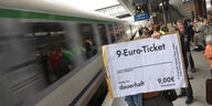 Ein Schild in Form einer Fahrkarte "9-Euro-Ticket dauerhaft" wird vor einem Zug in die Höhe gehalten