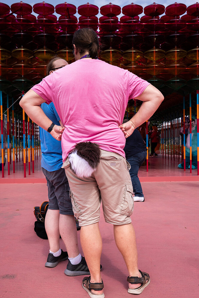 Zwei Menschen unterhalten sich, es ist der Rücken von einem Menschen zu sehen, der Hose und T-Shirt trägt - und einen Kostümschwanz