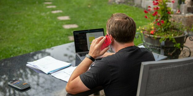 Ein Mann von hinten, telefoniert am Schreibtisch im Garten, vor sich ein Laptop.
