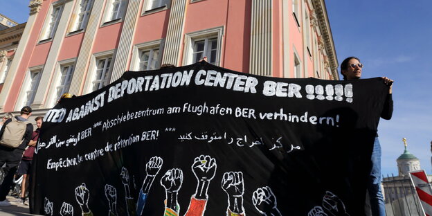 Vor dem Landtag in Potsdam demonstrieren Aktivist:innen gegen ein Abschiebezentrum am Flughafen BER, sie zeigen ein Banner.