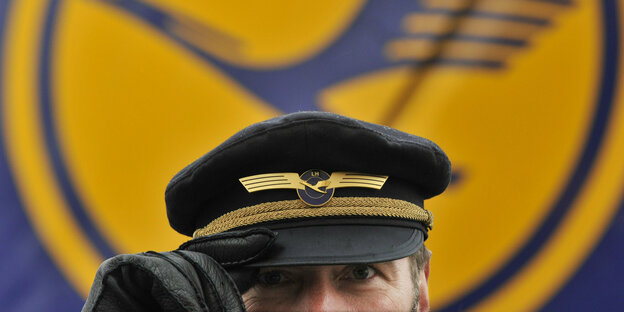 Mit seiner Uniformmütze auf dem Kopf steht ein Pilot der Lufthansa am Flughafen in Frankfurt am Main vor dem Lufthansa-Logo