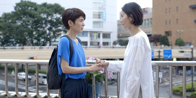 Zwei junge japanische Frauen stehen an einer Brücke in einer Stadt, halten sich die Hände und schauen sich in die Augen