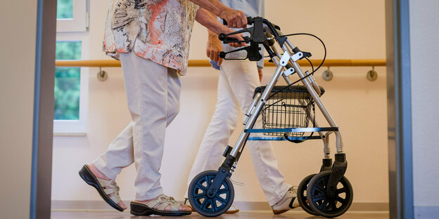 A woman with a walker rolls down a hallway, behind her a nurse