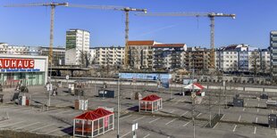 Ein großer, leerer Parkplatz aus Vogelpersektive. Auf dem Parkplatz befinden sich einige Häuschen für Einkaufswägen. Links ist ein Gebäude zu sehen und der Schriftzug "BAUHAUS", abgeschnitten zu erahnen. Hinter dem Parkplatz befinden sich Wohnblöcke und Kräne in Aktion.