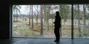 KZ Bergen-Belsen: Eine Person blickt aus einem dunklen Raum im Dokumentationszentrum durch ein großes Fenster auf die Gedenkstätte.